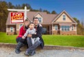 Family sitting on the curb in front of a house with a sold sign in the yard.